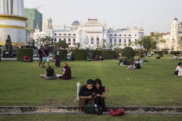 A couple look at a mobile device while other visitors and tourists sit in Maha Bandoola Garden as Yangon City Hall stands in the background, in Yangon, Myanmar, on Thursday, Feb. 4, 2016. Aung San Suu Kyi's party assumed control of the nation's parliament earlier this month as more than half a century of military rule in the country comes to an end. Photographer: Taylor Weidman/Bloomberg via Getty Images