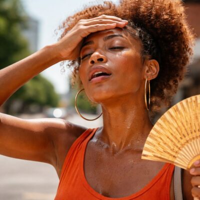 An AI-generated visual of a woman in a bright orange top sitting by a window, holding a glass of water to stay hydrated during the March 2026 Nigerian heatwave.