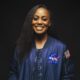 Dr Wendy Okolo, the first Black woman to earn a Ph.D. in Aerospace Engineering, wearing a blue NASA bomber jacket and smiling against a dark background.