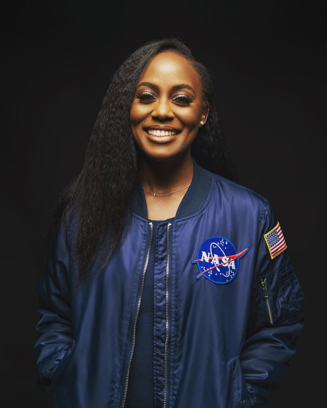 Dr Wendy Okolo, the first Black woman to earn a Ph.D. in Aerospace Engineering, wearing a blue NASA bomber jacket and smiling against a dark background.