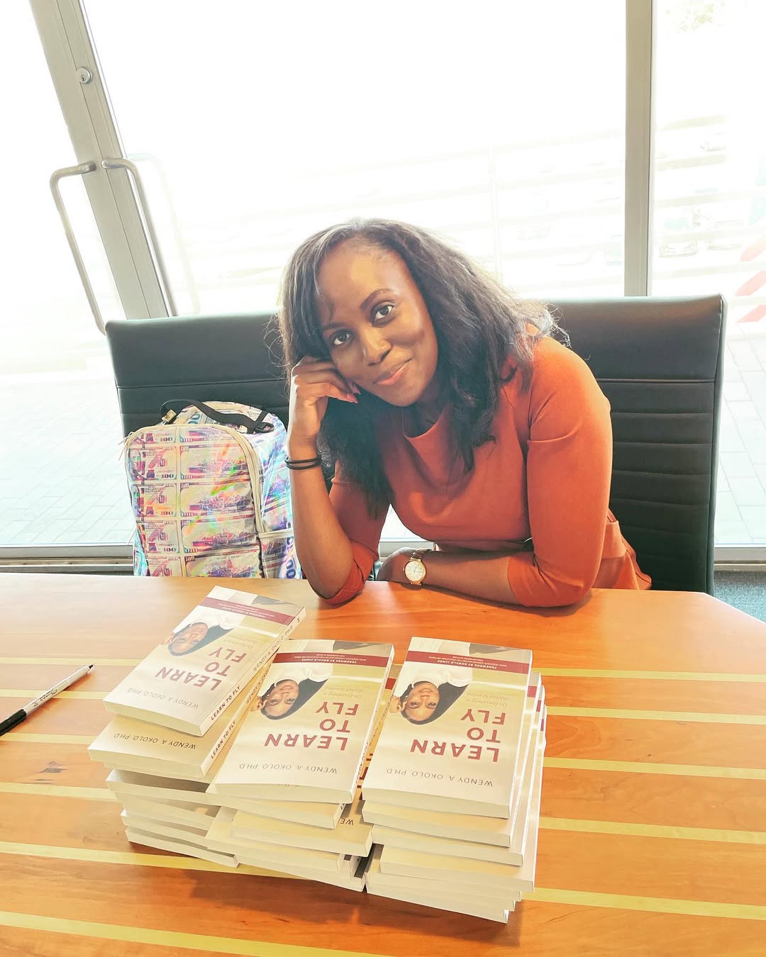 Dr Wendy Okolo sitting at a wooden table with stacks of her book "Learn to Fly," wearing an orange top and smiling for the camera at a book signing event.