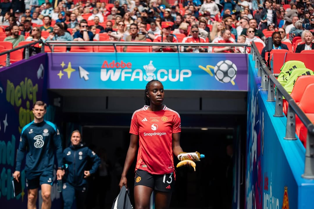 Manchester United midfielder Simi Awujo (number 13) emerging from the Kingsmeadow tunnel for the Women's FA Cup, clutching her boots and pre-match nutrition, with the Adobe WFA Cup branding and a sea of fans in United red behind her.