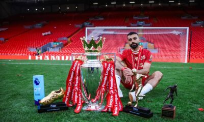 Mohamed Salah posing with the Premier League trophy, Golden Boot, and Playmaker award on the Anfield pitch.