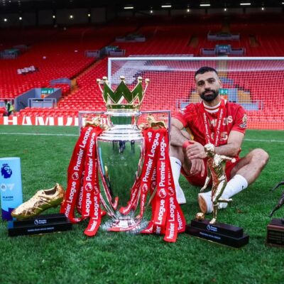 Mohamed Salah posing with the Premier League trophy, Golden Boot, and Playmaker award on the Anfield pitch.