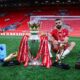 Mohamed Salah posing with the Premier League trophy, Golden Boot, and Playmaker award on the Anfield pitch.