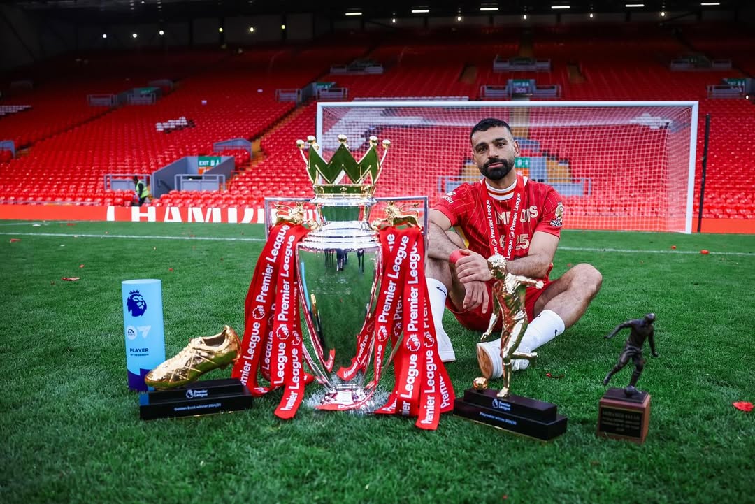 Mohamed Salah posing with the Premier League trophy, Golden Boot, and Playmaker award on the Anfield pitch.