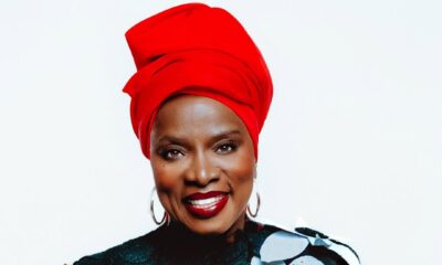 Close-up studio portrait of Angélique Kidjo wearing a red headscarf and a black top with colourful 3D floral appliqués, smiling against a white background for her "Hope!!" album campaign.