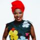 Close-up studio portrait of Angélique Kidjo wearing a red headscarf and a black top with colourful 3D floral appliqués, smiling against a white background for her "Hope!!" album campaign.