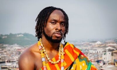 Ghanaian musician Fuse ODG draped in traditional Kente cloth and gold beaded necklaces, seated overlooking a coastal Ghanaian city.