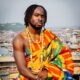 Ghanaian musician Fuse ODG draped in traditional Kente cloth and gold beaded necklaces, seated overlooking a coastal Ghanaian city.