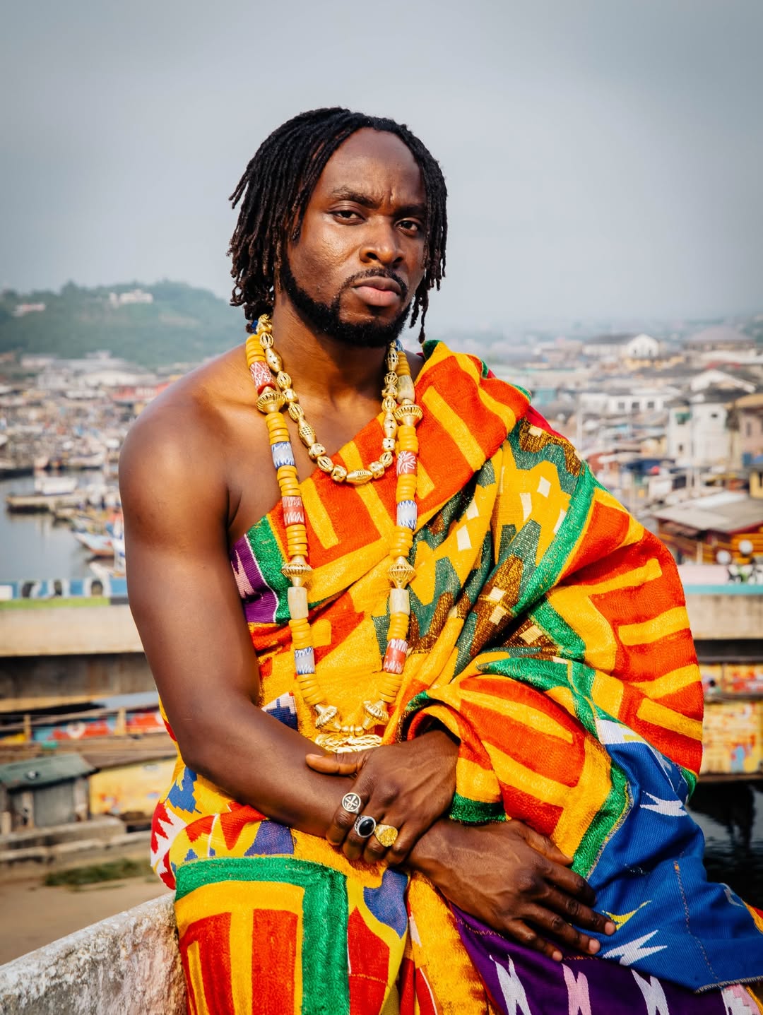 Ghanaian musician Fuse ODG draped in traditional Kente cloth and gold beaded necklaces, seated overlooking a coastal Ghanaian city.
