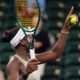 A close-up of Victoria Mboko in a white visor and tan tennis dress, holding her racket and a tennis ball while looking up during her victory over Amanda Anisimova at Indian Wells 2026.