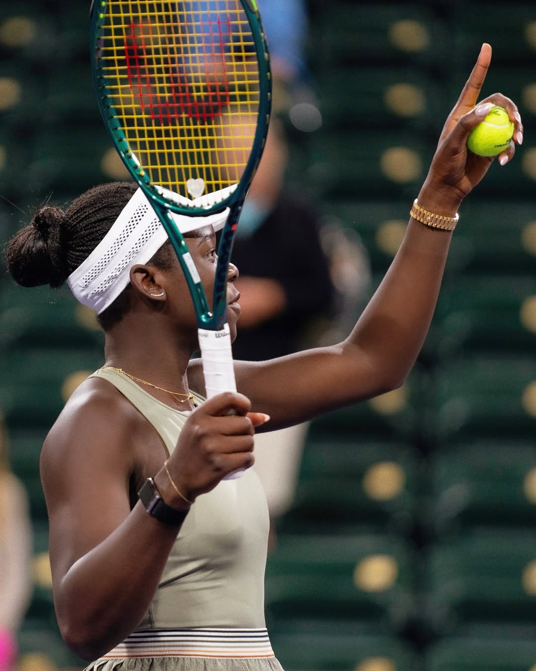 A close-up of Victoria Mboko in a white visor and tan tennis dress, holding her racket and a tennis ball while looking up during her victory over Amanda Anisimova at Indian Wells 2026.