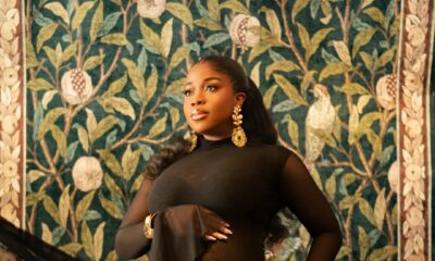 Close-up of fashion designer Veekee James in a black sheer mesh gown, featuring a gold letter charm belt and gold earrings.