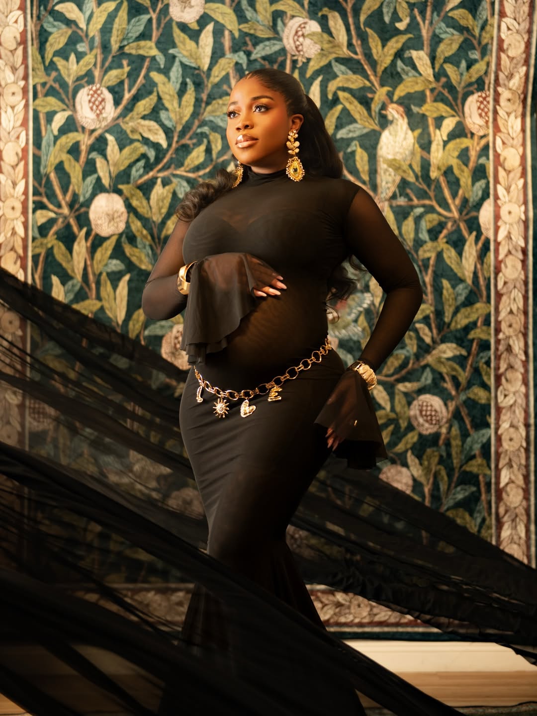 Close-up of fashion designer Veekee James in a black sheer mesh gown, featuring a gold letter charm belt and gold earrings.