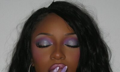 close-up of Ayra Starr with her eyes closed, showcasing a dramatic purple eyeshadow look with silver glitter, paired with glossy lilac lips and matching lilac nails.