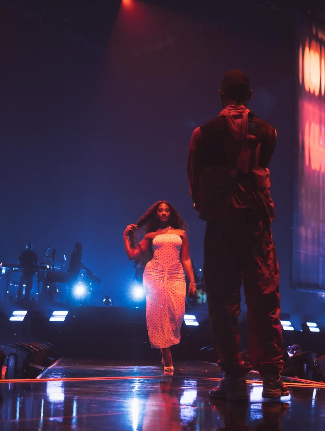 Tems walking toward Dave on a dark stage during the London stop of the Boy Who Played the Harp Tour. Tems is wearing a ruched polka dot dress while Dave stands in the foreground with his back to the camera.