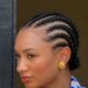 A profile close-up of Temiloluwa Ajibade in Accra, showing her neat, back-braided cornrows, gold ribbed stud earrings, and a blue dalmatian-print collar.