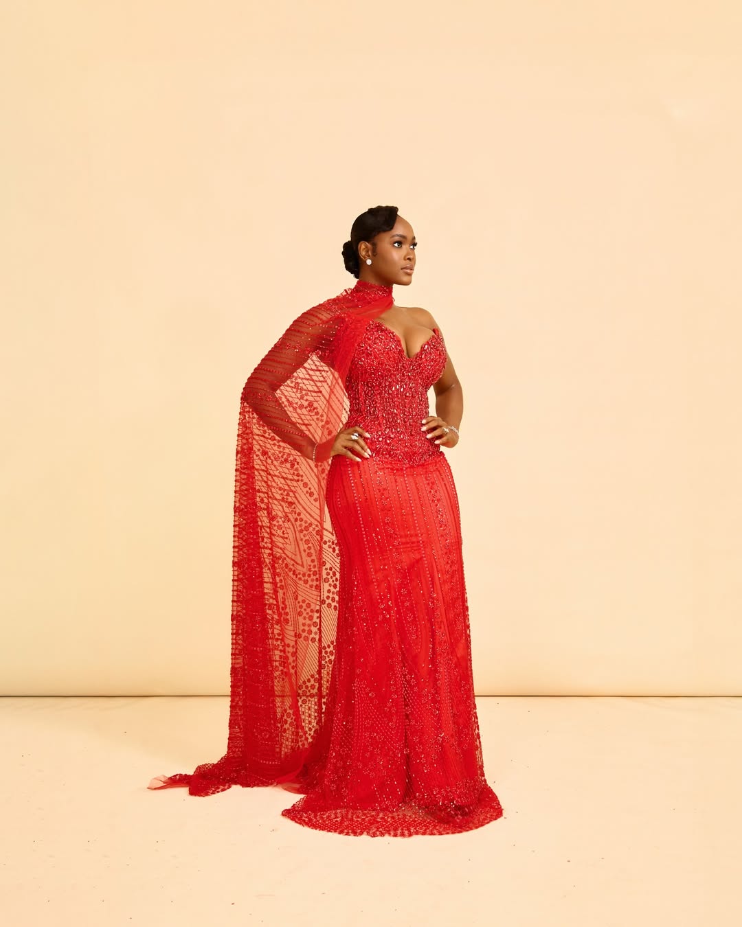 Nollywood actress Angel Anosike posing in a floor-length, embellished crimson red gown with a sheer dramatic cape for the Aba Blues movie premiere.