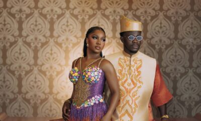 Simi Sanya and Femi Dapson posing against a patterned wallpaper backdrop in coordinated traditional after-party attire. Simi wears a purple gradient fringe mini dress with circular 3D appliqués while Femi wears a burnt-orange brocade ensemble with a gold-embroidered cream vest.