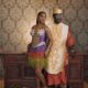 Simi Sanya and Femi Dapson posing against a patterned wallpaper backdrop in coordinated traditional after-party attire. Simi wears a purple gradient fringe mini dress with circular 3D appliqués while Femi wears a burnt-orange brocade ensemble with a gold-embroidered cream vest.