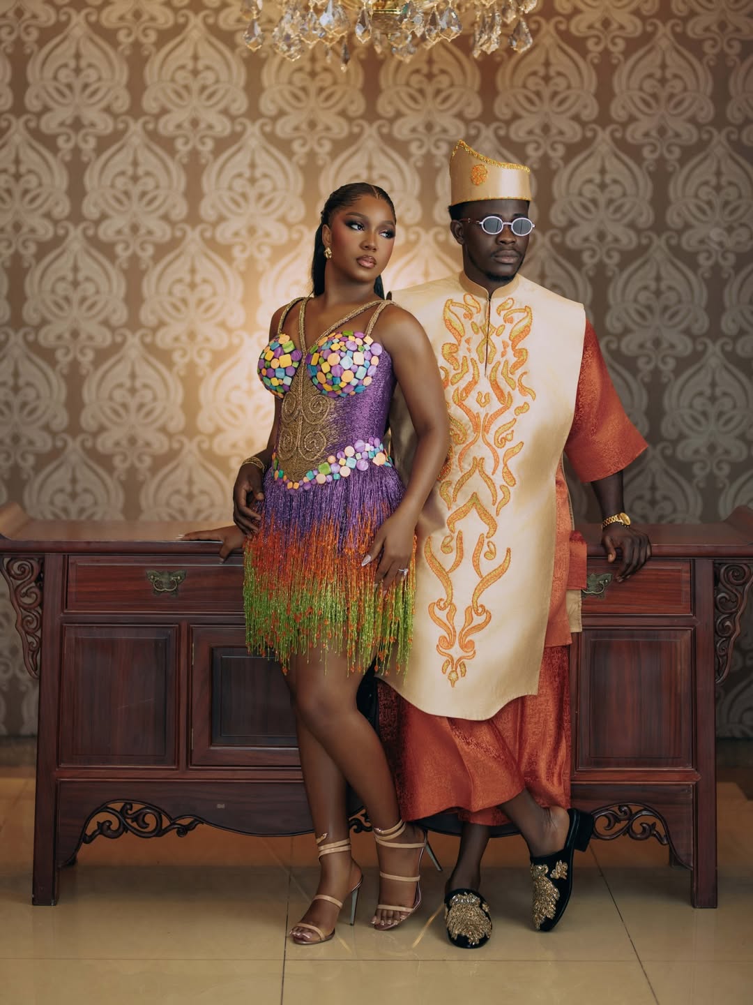 Simi Sanya and Femi Dapson posing against a patterned wallpaper backdrop in coordinated traditional after-party attire. Simi wears a purple gradient fringe mini dress with circular 3D appliqués while Femi wears a burnt-orange brocade ensemble with a gold-embroidered cream vest.