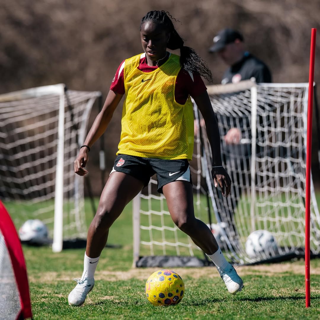 Midfielder Simi Awujo in deep focus during a Canada Soccer training session, wearing a maroon training top with a yellow mesh bib and black Canada-branded shorts, skillfully controlling a yellow and blue football on a grass pitch.