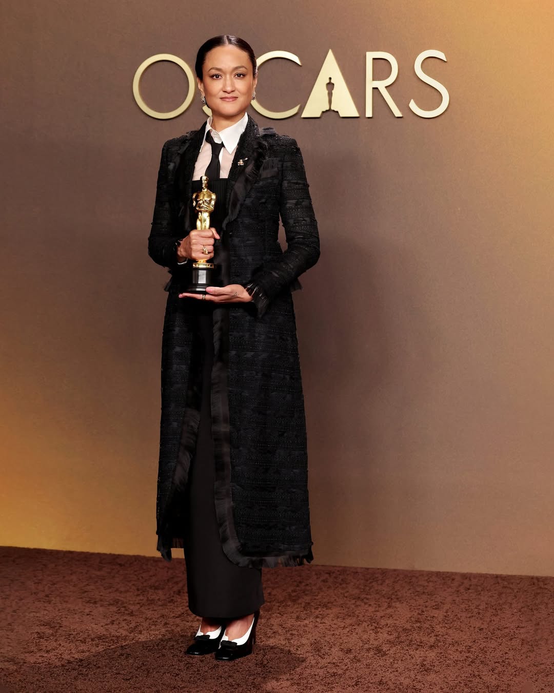 Cinematographer Autumn Durald Arkapaw holding her Academy Award for Best Cinematography for the film Sinners, wearing a black structured coat and tie at the 98th Oscars.