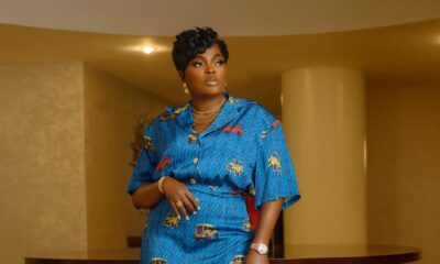 Funke Akindele in a teal blue Wanni Fuga two-piece set featuring a short-sleeved shirt and maxi skirt with a red and yellow vehicle print. She is accessorised with gold jewellery and has a short textured pixie haircut.