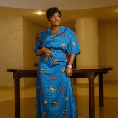 Funke Akindele in a teal blue Wanni Fuga two-piece set featuring a short-sleeved shirt and maxi skirt with a red and yellow vehicle print. She is accessorised with gold jewellery and has a short textured pixie haircut.