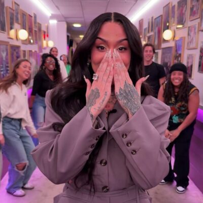 R&B singer Kehlani dancing through the Spirit Tunnel on The Jennifer Hudson Show. She is wearing a grey oversized belted blazer, a matching mini skirt, and burgundy tights, surrounded by the show's cheering crew.