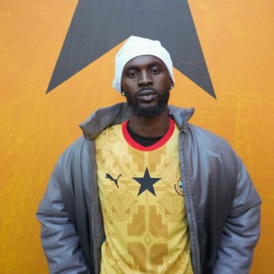 Ghanaian musician Black Sherif wearing the 2026 yellow Ghana away jersey with a grey jacket and white beanie.