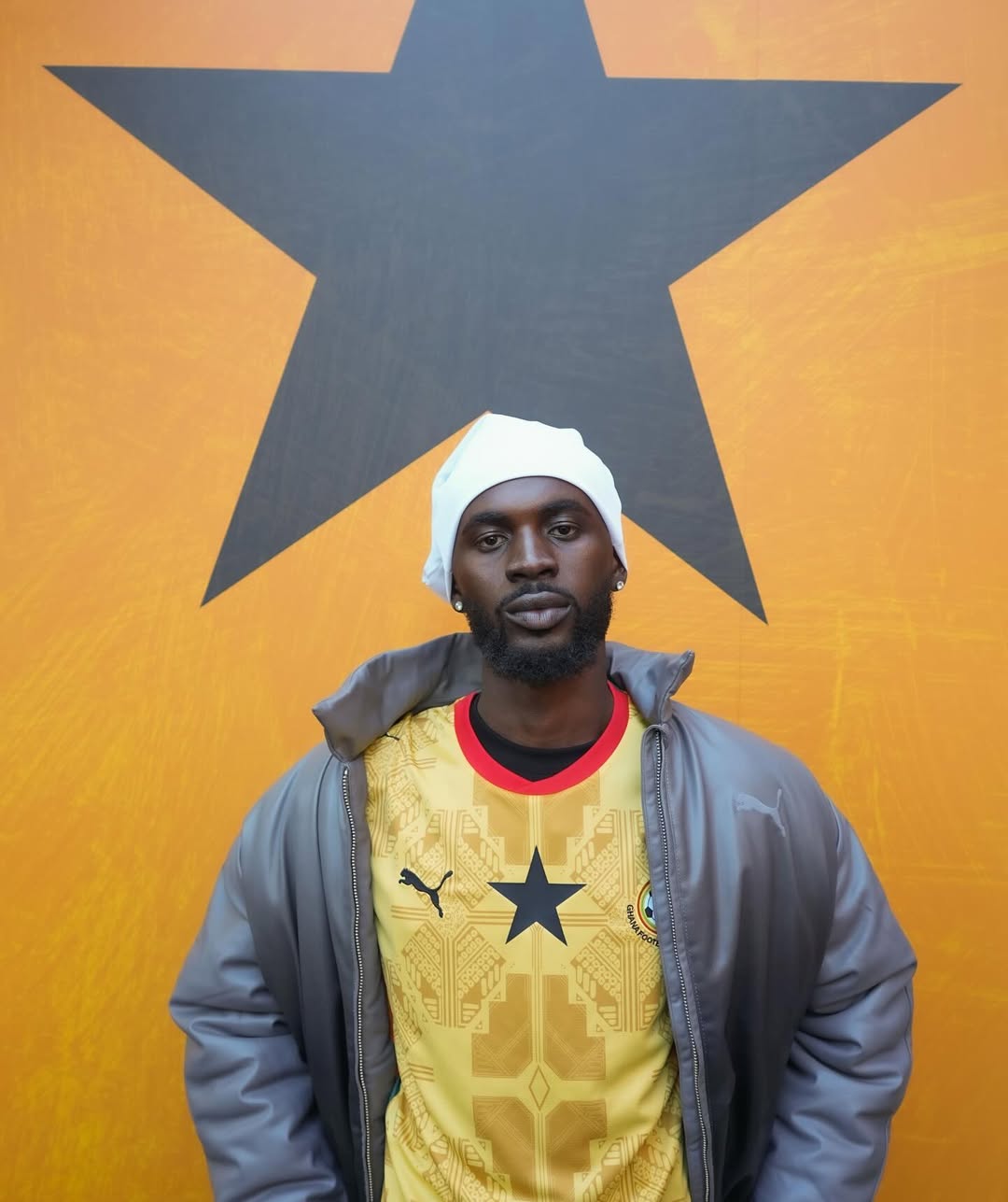 Ghanaian musician Black Sherif wearing the 2026 yellow Ghana away jersey with a grey jacket and white beanie.
