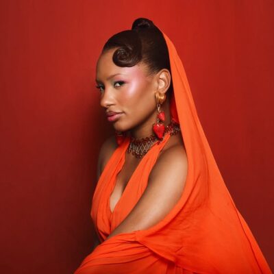 A close-up profile of Temi Ajibade (Otedola) featuring a vintage-style curled ponytail, glowing warm makeup, and statement gold earrings with red strawberry charms. She is draped in a vibrant orange fabric against a rich red background.
