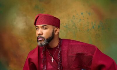 Close-up portrait of Banky W in a deep crimson agbada and fila holding a ceremonial Yoruba staff.