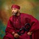 Close-up portrait of Banky W in a deep crimson agbada and fila holding a ceremonial Yoruba staff.