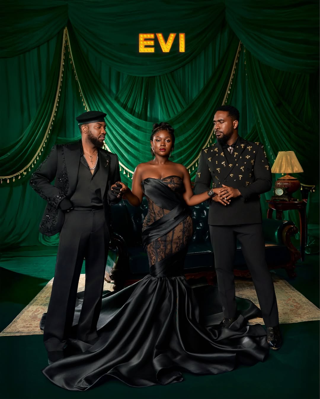 Osas Okonyon in a dramatic black strapless mermaid gown, positioned between co-stars Ibrahim Suleiman (left) and Uzor Arukwe (right) for a promotional portrait for the 2026 film Evi, directed by Uyoyou Adia. 