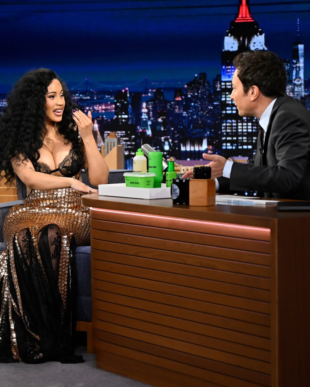 Cardi B seated at the desk with Jimmy Fallon on The Tonight Show, with her Grow-Good Beauty products displayed on a tray between them.