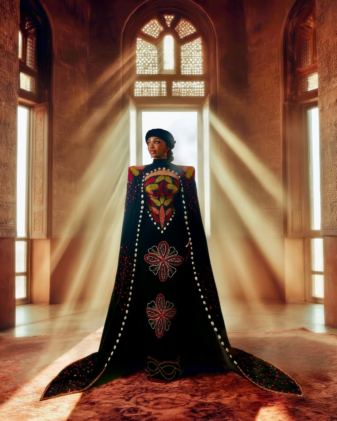 Chioma Ikokwu standing in a commanding black cape gown by Amy Aghomi. The cape is embroidered with bold botanical and geometric motifs in red, gold, and green, and grounded by a considered placement of cowrie shells lining the edges. She wears a black beret.