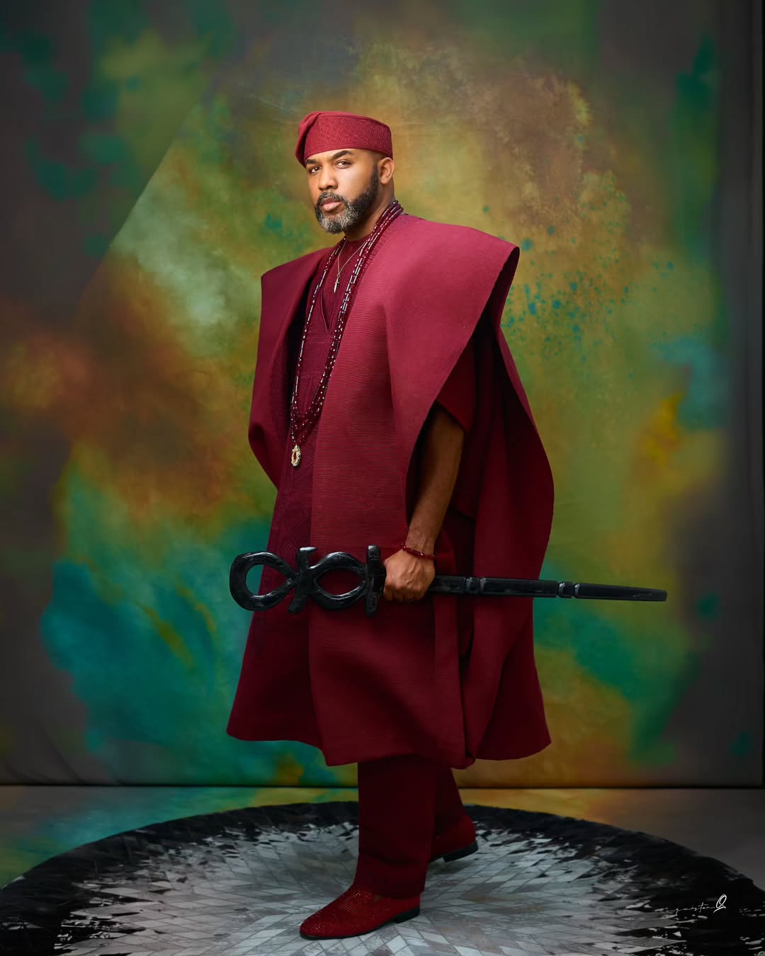 Full-length photo of Banky W in a structured crimson agbada and embellished red boots standing on a circular platform.