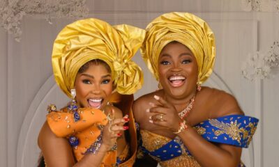 Funke Akindele and Iyabo Ojo laughing in coordinated blue and gold traditional lace outfits with elaborate yellow geles for the film Behind The Scenes.