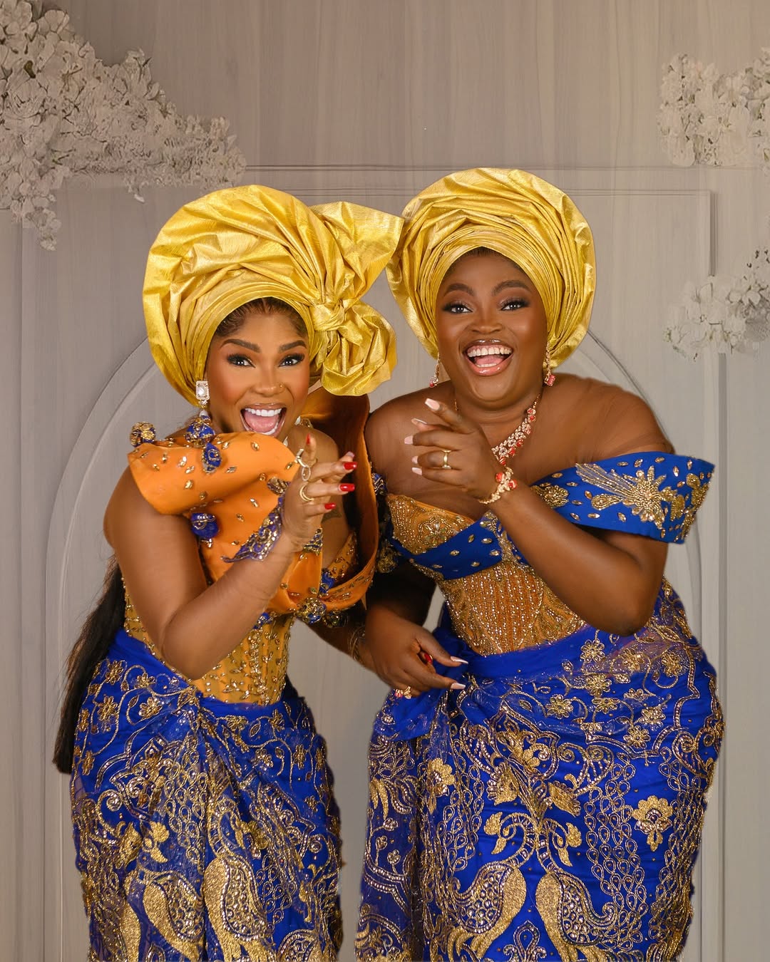 Funke Akindele and Iyabo Ojo laughing in coordinated blue and gold traditional lace outfits with elaborate yellow geles for the film Behind The Scenes.