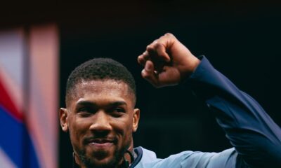 Anthony Joshua smiling and raising a fist while wearing an Under Armour tracksuit and a large Polymarket diamond chain.