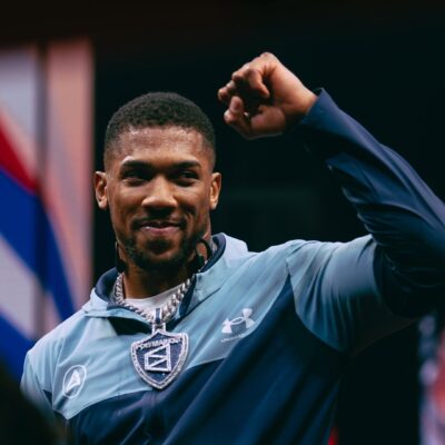 Anthony Joshua smiling and raising a fist while wearing an Under Armour tracksuit and a large Polymarket diamond chain.