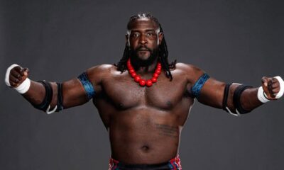 A studio portrait of Nigerian WWE wrestler Oba Femi, "The Ruler," standing in his signature wrestling gear with a red beaded necklace and blue armbands.