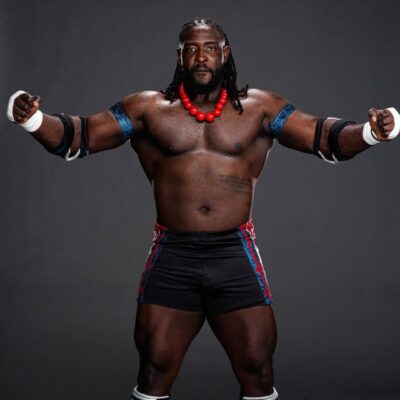 A studio portrait of Nigerian WWE wrestler Oba Femi, "The Ruler," standing in his signature wrestling gear with a red beaded necklace and blue armbands.