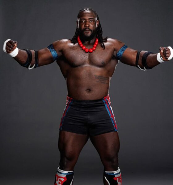 A studio portrait of Nigerian WWE wrestler Oba Femi, "The Ruler," standing in his signature wrestling gear with a red beaded necklace and blue armbands.