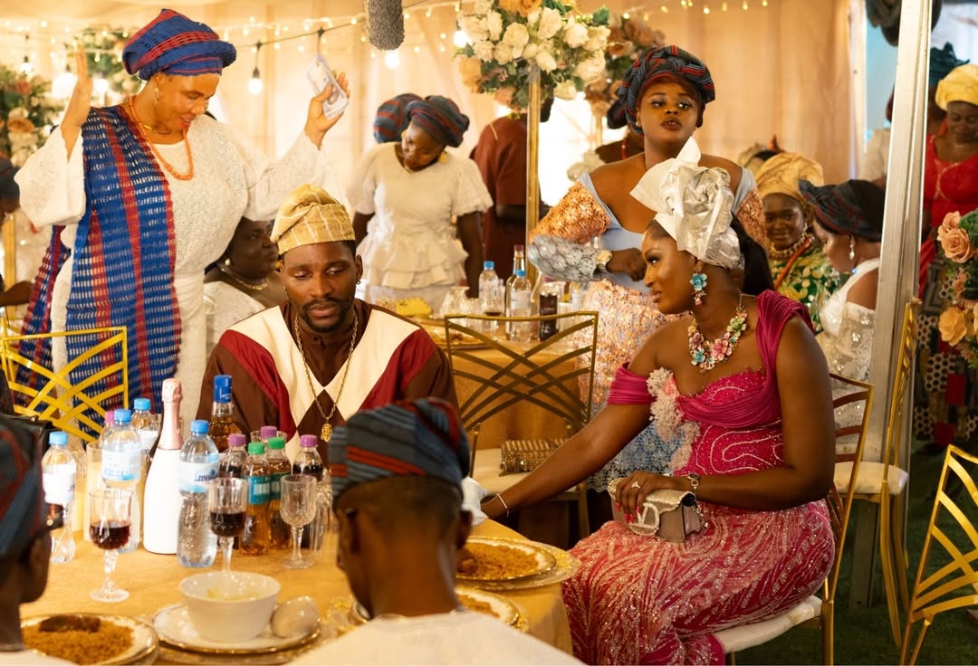 Scarlet Gomez in a bright pink lace traditional outfit and silver headgear (gele) seated at a wedding table next to Tobi Bakre on the set of the film Behind The Scenes.