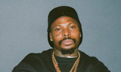 A studio portrait of Asake wearing a black sweatshirt, two gold Cuban link chains, and a black beanie, posing with his arms crossed against a neutral grey background.