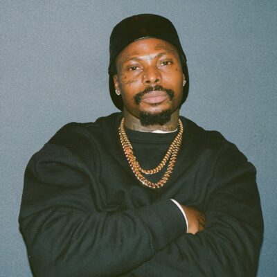 A studio portrait of Asake wearing a black sweatshirt, two gold Cuban link chains, and a black beanie, posing with his arms crossed against a neutral grey background.